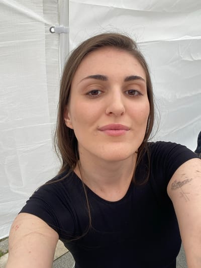 A selfie of a woman is taken outdoors, with a white tent-like structure in the background. The woman has long, brown hair and is wearing a black t-shirt. She has a small tattoo on her upper right arm, appearing to be a dandelion. There are a few red marks on her left arm, and some small red spots on her right arm. The background appears to be made of a white, textured fabric, possibly a tent or canopy, with some hardware visible. The lighting is bright and natural. The location is not explicitly visible in the image but is identified as Oktyabrskoye, Russia.