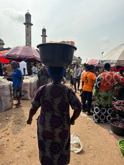 The image depicts a bustling outdoor market scene in Ogere, Nigeria, with a mosque visible in the background. In the immediate foreground, a person, viewed from behind, wears a long-sleeved purple dress patterned with white dotted floral designs and carries a dark-colored basin filled with light-colored goods, possibly packaged food items, balanced on their head, secured by a dark blue headwrap. The person's right hand is visible, clenched, holding a small white object.
To the left, a woman in a blue t-shirt and patterned skirt or shorts stands near a stack of large, white woven sacks, some labeled "FASCO," which appear to contain bulk goods like charcoal or grains. A red market umbrella provides shade overhead. Various individuals are visible in the mid-ground, moving between stalls. One person wears an orange t-shirt with a white number "5" on the back. Another individual, wearing a long green, orange, and black patterned dress, stands to the right, behind multiple black basins filled with red onions. A small green basket containing green produce is visible at the lower right edge. The ground is unpaved, reddish-brown earth, with some debris, including a clear plastic bag containing light material, scattered on the surface.
In the background, two prominent, light-grey minarets of a mosque, each topped with a small dome, rise against an overcast or hazy sky. Other indistinct structures are partially visible behind the market activity. The scene conveys active commerce under natural daylight.