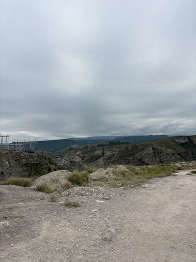 The image depicts a rugged, mountainous landscape with a cloudy sky. In the foreground, there is a dirt path with gravel and sparse vegetation. In the middle ground, the terrain becomes steeper, with rocky outcrops and greenery. To the left, a power substation with tall metal structures and electrical equipment is visible, situated on the edge of a canyon. The background features rolling hills and mountains stretching towards the horizon, under a heavily overcast sky. The scene suggests an industrial area integrated into a natural, remote environment, possibly near the Chirkey Dam in Chirkey, Russia.