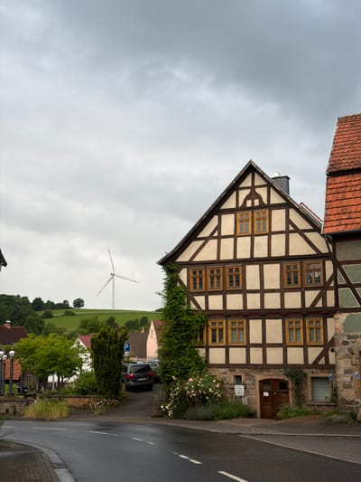 The image depicts a street scene in Ober-Gleen, Germany. In the foreground, a wet asphalt road curves to the left, with a brick sidewalk on the right. A building with a distinctive half-timbered facade dominates the right side of the frame. This building features brown wooden beams against a lighter colored infill, with multiple windows with yellow frames. At the base of this building, there is a wooden door and a window with blinds. To the left of this main building, a dense evergreen tree with blooming pink roses in front of it is visible. Behind this, a dark SUV is parked in a lot next to a small white building. Further in the background, a white wind turbine stands on a green, rolling hill under a cloudy, overcast sky. The landscape features scattered trees and other buildings with red tiled roofs. The overall impression is of a quiet village street on a damp day.
