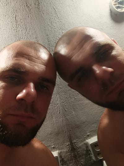 Two men are captured in a close-up, low-angle selfie, seemingly in a bathroom or similar space with textured, light-colored walls. The man on the left is in the foreground, with his face turned towards the camera. He has a shaved head, blue eyes, and a short beard. His skin appears slightly shiny, possibly due to sweat. To his right, the second man's head and shoulder are visible, also with a shaved head. His face is partially obscured and angled away from the camera. Behind them, the wall shows a distressed, peeling paint texture. To the far right, a white electrical outlet with two sockets is visible against the wall. The lighting is dim and cast from above, creating highlights on their heads and faces. The overall impression is informal and candid. There is no specific indication of the city of Pishchane, Ukraine, within the image itself.