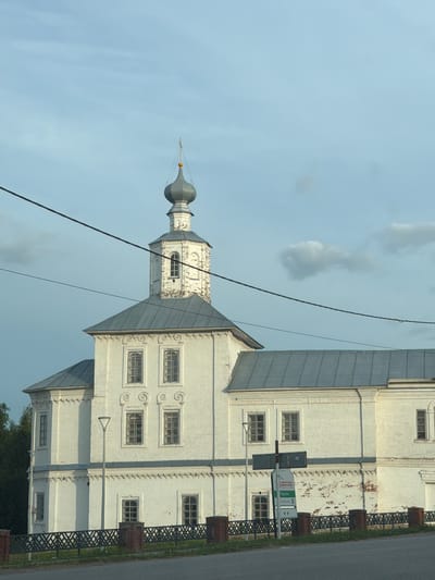 A white church with a grey roof and a prominent bell tower stands under a pale blue, cloudy sky. The church has multiple windows, some of which appear to be boarded up. A black metal fence with brick posts runs along the front of the property. A modern street lamp stands near the church. A sign with green and white lettering is visible in front of the building. Power lines are strung across the upper portion of the image. The scene appears to be an exterior view of a historic building in Cherdyn, Russia.