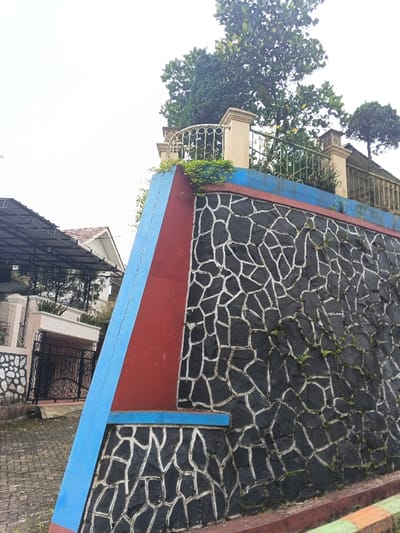 This image depicts an outdoor scene in Makilala, Philippines, under an overcast sky. The primary focus is a multi-section retaining wall. The main portion of the wall features a random rubble pattern composed of irregularly shaped dark grey or black stones set with light-colored mortar. Sparse green vegetation and moss are visible growing in the mortar lines. An angled, reddish-brown structural element is present on the left side of the stone wall, marked by a distinct vertical light blue stripe and a short horizontal light blue ledge at its base. A continuous light blue band runs along the top edge of the entire retaining wall. Above this band, a cream-colored decorative railing, featuring posts and balusters, is visible. Behind the railing, dense green foliage and several trees, including a jackfruit tree with visible fruit, are present. A smaller, manicured tree stands further to the right. To the left of the retaining wall, a paved surface leads to a black ornamental metal gate with intricate scrollwork. Beyond the gate, a white residential building with a dark-pitched roof and an adjacent structure with a dark, sloped canopy supported by columns are partially visible. A section of a multi-colored curb (orange, green, and brown stripes) is visible at the bottom right edge of the frame. No individuals are present in the scene.