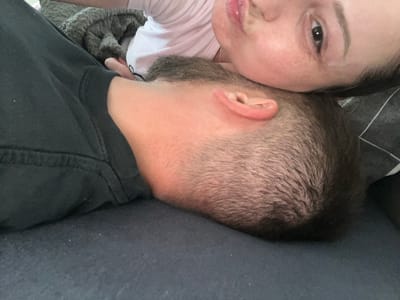 The scene, situated in Heusden-Zolder, Belgium, depicts two individuals in close proximity, lying indoors on a dark-colored, soft surface, likely a bed or couch. The lower half of the frame shows the back of a person's head and upper neck, characterized by short, dark hair and light-toned skin. This individual is wearing a dark, possibly black, garment, with a visible sleeve cuff in the foreground. Resting on or adjacent to this individual's head, the upper half of the frame features a second person, appearing female, whose face is visible. This individual displays open dark eyes looking towards the viewer, slightly pursed lips, light skin with freckles on the cheek, and defined eyebrows. They are clad in a light-colored, possibly white or light pink, top, and a thin silver chain is visible around their neck. A textured dark grey blanket is partially visible in the upper left background, and a dark pillow with a subtle geometric pattern is present in the upper right.