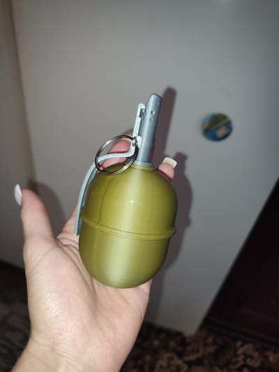 A person's hand holds a green, oval-shaped object with a silver lever mechanism and a ring on top, resembling a grenade. The background is a plain, lightly colored wall, with a blurry circular object, possibly a magnet or sticker, visible in the upper right corner. The person's fingernails are painted white. The location is the city of Voskobiinyky, Ukraine.