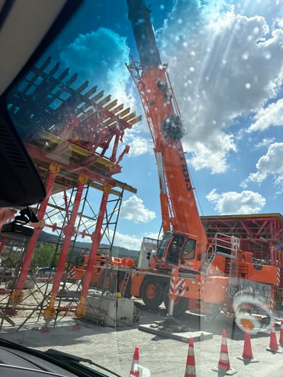 The image captures a construction site in the Municipal Unit of Polichni, Greece. A large orange crane dominates the scene, its boom extended upwards towards a bright blue sky filled with fluffy white clouds. The crane is positioned next to a substantial red and yellow scaffolding structure, likely part of a building or infrastructure project. In the foreground, the reflection on a window or windshield is visible, showing a dashcam mounted on the left. The ground is covered in gravel and dirt, with several orange and white traffic cones placed around the site. Some concrete blocks and rebar are also visible, indicating ongoing construction work. The sun is shining brightly, casting glare and lens flare on the image.