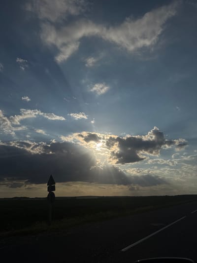 The image depicts a scenic view of a rural landscape in Азгінавіцкі сельскі Савет, Belarus, during what appears to be sunset or sunrise. The sky is filled with dramatic clouds, some dark and heavy, others illuminated by the sun which is partially obscured, creating visible sunbeams. In the foreground, a paved road runs diagonally from the bottom right towards the horizon, with a white line marking its edge. To the left of the road, a signpost stands in a field of dry grass. The signpost has two stacked, triangular signs, one above the other, facing the viewer, and a circular sign below them. Beyond the signpost, a dark, flat horizon is visible, with what seems to be agricultural fields and distant trees or structures. The overall impression is one of a serene, natural environment.