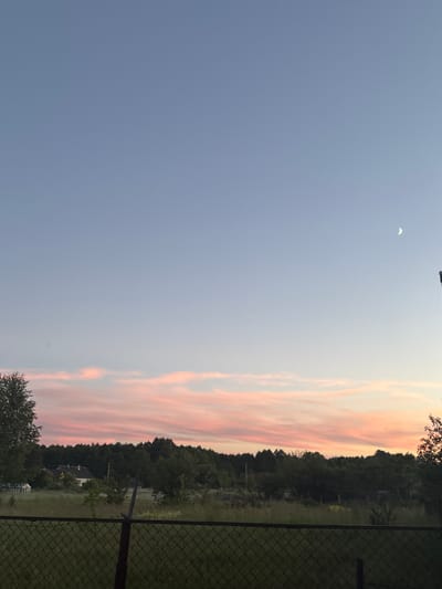 The image depicts a rural landscape at dusk, with a faint crescent moon visible in the pale blue sky. A gradient of pink and orange hues colors the horizon, hinting at a sunset. In the middle ground, a dense line of trees forms a natural backdrop to a few scattered houses with visible roofs. A chain-link fence runs across the foreground, partially obscuring a grassy field. The scene suggests a quiet evening in the countryside, likely located in the vicinity of Ганчарскі сельскі Савет, Belarus.