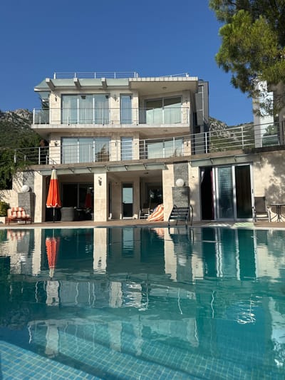 In Brca, Montenegro, a modern villa with a swimming pool is captured. The villa, constructed with light-colored stone, features multiple levels with balconies and large windows. A clear blue sky dominates the upper portion of the image. In the foreground, the turquoise water of a swimming pool reflects the villa and the surrounding landscape, which includes a lush green tree on the right and a mountainous terrain in the background. The pool's tiled bottom is visible through the clear water. Several outdoor furniture pieces, including lounge chairs and a table, are placed around the pool and on the villa's patio. An orange umbrella stands near a seating area. The scene is bathed in bright sunlight, creating sharp contrasts and reflections. There are no people visible in the image.