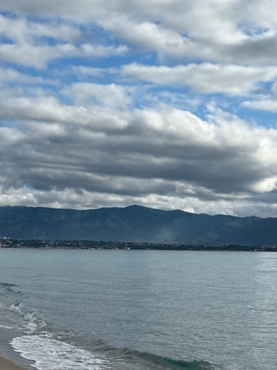 The image displays a coastal landscape in Quartu Sant'Elena, Italy. The foreground features a light-colored sandy beach on the lower left, where small, foamy waves break against the shore. The midground is dominated by calm, light blue-green sea water, extending across the frame. In the distance, a low-lying coastline is visible, characterized by dispersed light-colored structures and green vegetation. Behind this coastal strip, a dark, undulating mountain range stretches horizontally across the background. A distinct column of white smoke rises vertically from the distant coastline, positioned slightly right of the center. The sky is partially covered by a mix of stratocumulus clouds, presenting both dark gray and white formations, with intermittent patches of clear blue sky visible between the cloud layers. No individuals, specific vessels, or other significant objects are identifiable within the scene.
