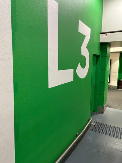 This image captures an indoor, utilitarian setting, likely within a parking facility or a service area, located in Indooroopilly, Australia. The most prominent feature is a bright green, textured floor, heavily painted with large, white alphanumeric characters and lines. The number "13" is clearly visible in white paint on the green floor, partially cropped, suggesting a designated parking spot or zone number. A thick white line runs along the bottom edge of the frame, possibly marking a boundary.

In the mid-ground and background, various architectural elements are visible. A grey wall with a distinctive perforated section runs along the right side, suggesting ventilation or a design feature. A thin, polished metal handrail is mounted horizontally along this wall. To the left, a raised platform or structure, also painted in the same vibrant green, extends across the scene. This structure includes a recessed rectangular panel or door with a small silver latch. Further back, a lighter-colored wall, possibly white or light grey, forms a corner with the green structure. A small, red-and-white sticker is affixed to this lighter wall, but its text is illegible. The scene is brightly lit by artificial light sources. No people or specific activities are depicted; the image presents a static view of the environment.