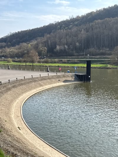 The image displays an outdoor scene in Mertert, Luxembourg, under daylight with a partly cloudy sky. The foreground features a curved, light grey concrete embankment defining the edge of a body of water with minor surface ripples. Adjacent to the embankment, an asphalt surface is delineated by a series of white posts connected by chains. Two larger grey bollards with yellow tops stand on the asphalt. A red lifebuoy is mounted on a stand near a prominent black, multi-tiered structure that projects into the water. This structure includes metal railings, a set of stairs descending towards the water, and a tall, slender black column topped with a light fixture. Several white placards are affixed to its sides. Across the water, a strip of green grass precedes a transportation corridor that contains a road with multiple vehicles (several cars and at least one flatbed truck) and a railway line with overhead electrification infrastructure. The background is dominated by a steeply rising, densely forested hillside, primarily composed of bare deciduous trees with some interspersed evergreens. No people are visible.