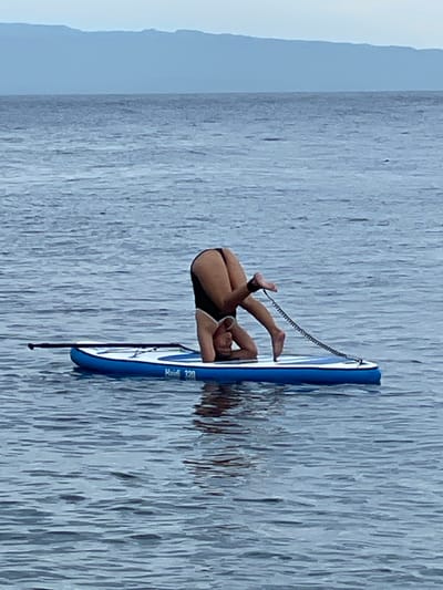 A person performs a headstand on a paddleboard in the calm waters off Dauin, Philippines. The person is wearing dark-colored swimwear and has their legs extended upwards, balanced on their head and forearms. A coiled leash connects their ankle to the paddleboard. The paddleboard is blue and white, with a visible model number "H-320". The background features a hazy blue mountain range against a pale sky. The water is calm with slight ripples.