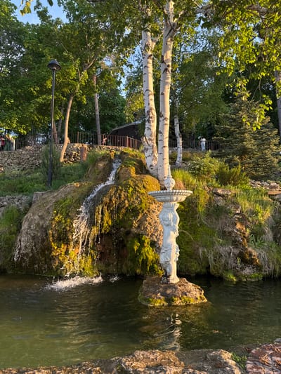 The image depicts a waterfall feature in Krasny Klyuch, Russia. Water cascades over moss-covered rocks into a pool. Atop the rocks, a white statue sits on a tiered platform. Birch trees stand adjacent to the rocks. To the left of the waterfall, there is a stone wall and a lamp post. A set of stairs leading upwards is visible in the background. The environment includes lush greenery and trees surrounding the waterfall.