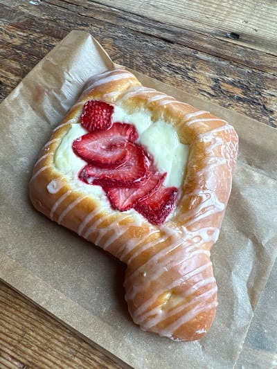 A strawberry danish with white icing is displayed on a piece of brown parchment paper, which rests on a wooden surface. The danish is golden brown with a creamy white filling and sliced strawberries on top. The icing is drizzled in a pattern over the pastry. The wooden surface appears to be a tabletop with visible grain and texture. The image is taken from a top-down perspective. The scene is located in Long Beach, United States of America.
