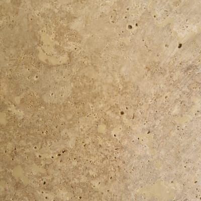 The image is a close-up, abstract view of a textured surface. The dominant color is a light tan or beige, with darker brown and lighter cream-colored variations creating a mottled and uneven appearance. There are numerous small, dark circular indentations or holes scattered across the surface, giving it a porous or rough texture. Some areas show faint striations or lines, suggesting a natural material like stone or wood with a weathered or rough finish. The lighting appears to be diffuse, casting no harsh shadows and highlighting the texture. There are no discernible objects, people, or a specific scene visible. The image is purely textural and lacks any contextual clues about time of day, weather, or location, other than being abstract. No text is visible in the image.