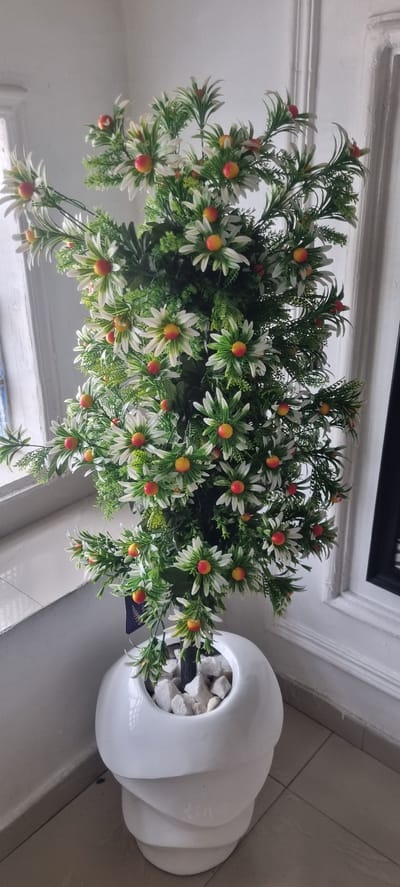 The central subject is a decorative potted plant, likely artificial, featuring dense green foliage. Numerous daisy-like flowers with white petals, fading to green towards their centers, are interspersed throughout the plant. Each flower has a prominent, spherical red-orange center, resembling a small berry or fruit.
The plant is housed in a modern, glossy white ceramic pot with a distinctive layered or sculptural design. The pot's opening is filled with small, irregularly shaped white decorative stones or pebbles.
The setting appears to be indoors, with light-colored (possibly white or light grey) walls and floor tiles forming the background. A dark horizontal element, possibly part of a window frame or furniture, is visible in the upper right portion of the image, along with a white architectural trim on the wall.
No activity or event is taking place; the image is a still life capturing the plant as a decorative object.
Notable details include the bright, even lighting, suggesting an indoor environment. The photograph is taken from a slightly elevated perspective, looking down into the plant. A small, dark-colored tag or label is partially visible near the base of the plant, but any text on it is illegible. The provided location context for this media file is Alasia, Nigeria, although no specific visual cues within the image confirm this.