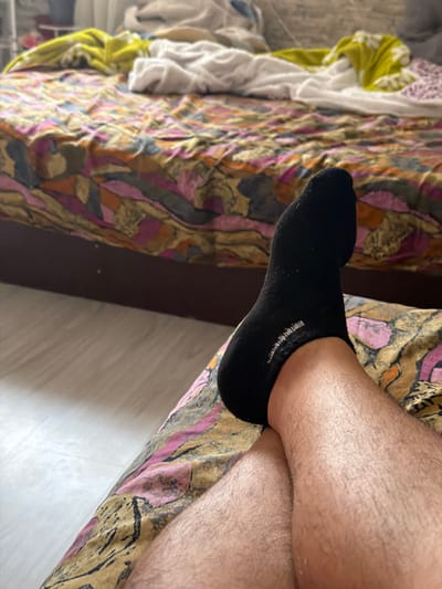 The image depicts an indoor, residential scene, likely a bedroom, viewed from a low angle. In the foreground, the lower legs and one foot of an individual are prominently displayed. The legs are bare, covered in hair, and crossed at the ankles. The left foot (from the viewer's perspective) is clad in a black, low-cut sock, which exhibits white lettering or a logo on its lateral side. The individual's legs are resting on a piece of bedding characterized by a multicolored, abstract pattern predominantly featuring shades of pink, orange, brown, grey, and gold. Behind the legs, a bed with a dark brown wooden frame is visible. This bed is covered with similar patterned bedding, which appears disheveled, with white and yellow fabric (likely sheets or blankets) crumpled on its surface. To the immediate left of the bed, a light-colored floor, resembling laminate or light wood, is visible. The specific location within Kermen, Bulgaria, cannot be visually confirmed from the elements present in the image.