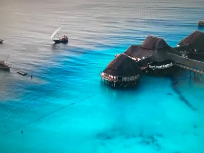 The image shows an aerial view of a tropical coastal scene, likely a resort area with buildings on stilts over clear, turquoise water. A group of thatched-roof buildings is connected by a wooden pier. Several boats are visible on the water, including one sailboat. The water clarity suggests a shallow, sandy bottom. Two individuals can be seen swimming. The scene does not appear to be in Basingstoke and Deane, United Kingdom.
