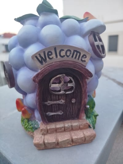 This is a close-up, eye-level shot of a whimsical, grape-shaped fairy house. The house is predominantly light purple, with round, bulbous sections mimicking grapes. Dark green leaves sprout from the top. A rustic wooden door with metal hinges and a circular window is featured prominently on the front. Above the door, a curved sign reads "Welcome" in black, hand-painted lettering. There are two small, circular windows on the side of the house, one of which is partially visible. The house is adorned with small, colorful mushroom-like figures and green foliage at its base, suggesting a garden setting. The house is placed on a flat, gray surface, possibly a ledge or windowsill. In the background, blurred buildings with windows are visible, hinting at an outdoor urban or suburban environment. The lighting suggests it is daytime. The overall impression is one of a charming, miniature dwelling designed to evoke a sense of fantasy and hospitality.
