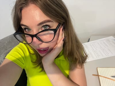 A medium shot shows a woman with shoulder-length brown hair, wearing a bright neon yellow ribbed short-sleeved shirt and black cat-eye glasses. She is indoors, likely in an office or study, as evidenced by papers and a notebook on a desk. Her left hand is resting on her cheek, with her tongue playfully peeking out from her lips, which are coated in sparkly pink lipstick. Her green eyes are looking directly at the camera. The background includes a grey upholstered chair on the left and a desk with documents and a notebook on the right. The lighting is bright, creating some shine on her skin.
