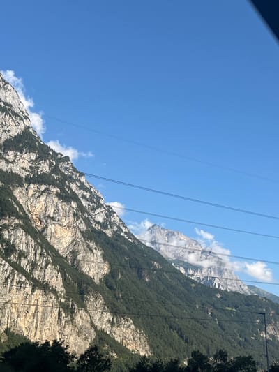 The image displays a mountainous landscape near Schattdorf, Switzerland, characterized by steep, rocky formations and dense coniferous forestation under a clear blue sky with scattered white clouds. A prominent mountain face with visible rock striations and green tree coverage occupies the left and center of the frame. In the distance to the right, a higher, more rugged peak with light-colored rock, possibly snow-dusted, is partially shrouded by clouds. Multiple dark, parallel power lines traverse the mid-section of the image horizontally. Dark green foliage, likely tree tops, is visible along the bottom edge of the frame. No individuals, specific actions, or additional discernible man-made structures are present.