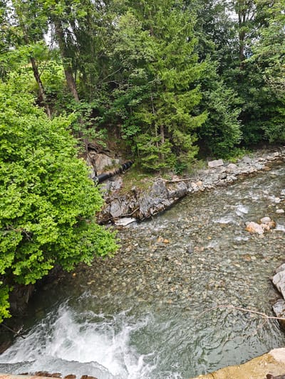 The image depicts a fast-flowing river with a rocky bed and bank, surrounded by lush green trees and vegetation. In the upper left portion of the image, there is a cluster of dense green foliage. A black pipe is visible protruding from a rocky incline on the left bank of the river, partially obscured by trees. The water is clear, revealing the various sized stones and pebbles on the riverbed. The scene appears to be a natural, outdoor setting, likely a forested area near a body of water. The specific location in the city of Filzen, Austria is not determinable from the visual information provided in the image itself.
