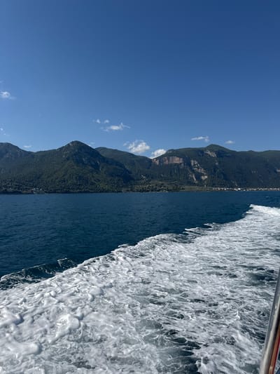 An eye-level, wide shot captures the deep blue waters of a body of water in Stroggili, Greece, with a foamy white wake trailing from the right side of the frame. Lush green mountains rise in the background, dotted with small white structures that suggest a settlement along the shoreline. The sky above is a clear, vibrant blue, with a few wispy white clouds scattered across it. The image gives the impression of a journey by boat on a bright, sunny day.