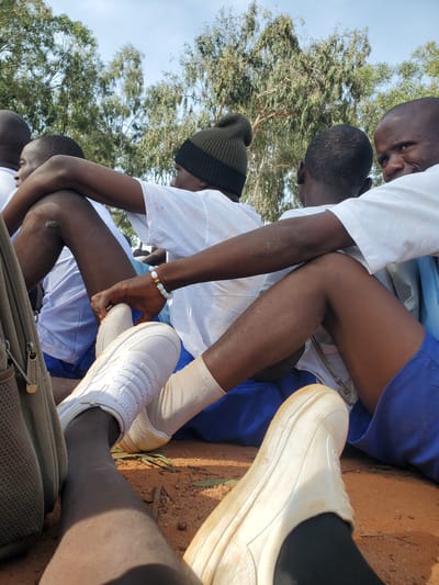 In Barakin Rafin Gora, Nigeria, a group of young people, likely students, are seated outdoors. They appear to be gathered in a relaxed, informal setting, possibly during a break or for an event. The individuals are seen from the waist down, with their legs crossed and arms resting on each other, suggesting a sense of camaraderie.

The individuals are wearing school uniforms, consisting of white shirts and blue shorts or skirts. Some have on white sneakers and socks. One person is wearing a dark knit hat. In the foreground, a backpack with a zipper is visible.

The ground is covered in reddish-brown dirt, with some dry leaves scattered around. In the background, several trees with green foliage are visible against a light blue sky. The overall impression is one of a bright, outdoor environment, typical of a school or community gathering space in Nigeria.