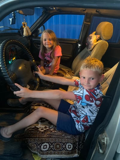 The image depicts the interior of a car, likely in the village of Yandyki, Russia. Two children are present. A young girl with blonde hair, wearing a pink t-shirt, is seated in the passenger seat, looking towards the camera. To her right, a young boy with blonde hair and wearing a Spider-Man themed shirt and blue shorts is seated in the driver's seat, with his hands on the steering wheel. The car's interior is visible, including the dashboard, steering wheel, seats, and seatbelts. A decorative item, possibly a stuffed animal, hangs from the rearview mirror. The background outside the car windows shows what appears to be a blue, corrugated metal structure, possibly a shipping container or building. The lighting suggests it may be dusk or evening.