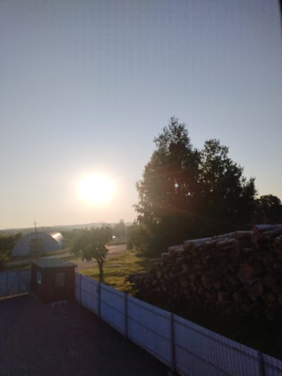 This is a photograph taken at dusk in the Вароніцкі сельскі Савет, Belarus. The scene depicts a rural landscape with a clear sky. In the foreground, there is a dark asphalt surface with a small red building and a light-colored fence. To the right of the fence, a large pile of cut logs is visible. In the middle ground, trees are scattered across a grassy area, with a large greenhouse-like structure on the left. The setting sun, a bright, luminous orb, is visible in the upper left portion of the image, casting a warm glow over the landscape. The overall atmosphere suggests a peaceful, rural setting at the end of the day.
