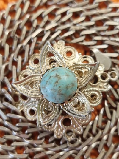 The image displays a close-up, overhead view of a piece of ornate jewelry, possibly a brooch or a pendant, set against a textured, orange background. The jewelry features a central, oval-shaped, light blue stone with visible matrix patterns, likely turquoise. This stone is surrounded by a decorative silver-toned metal setting. The setting has a floral or star-like design with multiple intricate, layered petals or filigree elements. The metalwork appears to be detailed, with small raised dots and swirling patterns within the filigree. The entire piece is partially overlaid with a silver-colored chain, which has wide, flattened links and is arranged in a loose, overlapping pattern. The chain's texture is prominent, with individual links clearly defined. The lighting is diffused, creating soft shadows and highlighting the metallic sheen of the chain and the texture of the stone. There are no people or visible actions. The scene is static, focusing solely on the details of the jewelry and chain.