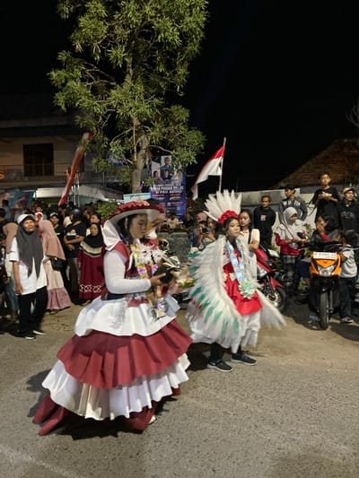 The image depicts an outdoor night scene in the city of Kedungbanteng, Indonesia, showing a public gathering or parade. In the foreground, two individuals in elaborate costumes are featured. The person on the left wears a multi-tiered gown with alternating red and white layers, a wide-brimmed red hat, and a white long-sleeved top; they are holding a colorful garland and a small object. The individual on the right is dressed in a red costume with large white and green feathered wings, a white feathered headpiece adorned with red flowers, and a long white garment, also wearing a colorful garland. Both are standing on a paved surface.

In the midground and background, numerous spectators, consisting of adults and youths, are present on both sides, observing the scene. Several individuals are standing, and some appear to be seated. Parked on the right side are at least two motorbikes, one red and one black and orange.

The environment includes a prominent tree with green foliage in the center-left. Buildings are visible in the background, including a two-story structure on the left. Two Indonesian flags (red and white) are displayed on poles. A large banner in the background features images of people and text, including "HAPPY FESTIVAL CARNAVAL 2023" and "H PRO AUDIO." The scene is illuminated by artificial light sources against a dark night sky.