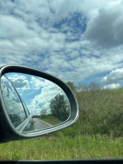 This image, taken in Pishurka, Bulgaria, captures a perspective from inside a vehicle, primarily focusing on the car's right-hand side mirror and the surrounding environment. The image is oriented with the sky on the left and the ground on the right, suggesting a rotation from the original capture.

The main subjects include a prominent, dark-framed car side mirror in the lower central and right portion of the frame. The background on the left is dominated by a bright blue sky filled with scattered white and light gray cumulus clouds. To the right, outside the car, a lush green landscape with trees and grass is visible, suggesting a roadside or rural setting.

The mirror's reflective surface clearly displays a panoramic view of the sky and clouds, mirroring the conditions outside. Within this reflection, a paved road can be seen receding into the distance, bordered by trees and vegetation, indicating that the car is in motion. Parts of the vehicle's own structure are also visible in the lower section of the mirror's reflection.

The scene depicts a daytime setting under fair, partly cloudy weather conditions. The immediate surroundings outside the car suggest a rural or semi-rural road environment. There are no people visible, nor is any text present in the image.