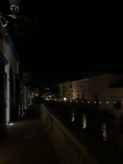 The image depicts a nighttime scene at Plaza Machado in Mazatlán, Mexico. Along the right side of the frame is a building with decorative trim and a sign on its wall. Small lights illuminate a pedestrian walkway. Tables and chairs are present along the walkway, suggesting outdoor dining areas. The left side of the image is predominantly dark, possibly showing a lack of buildings or structures. In the foreground, elements of a building or structure are visible, with some equipment attached to it. The overall lighting is dim, and the image is grainy. There is no apparent presence of people.