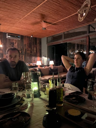 A photo, likely taken at night in Livingstone, Zambia, shows a group dining indoors, oriented vertically. A man with salt and pepper hair and a dark t-shirt is at the table's edge looking at the camera with an intense facial expression. Across the table, a woman in a dark top has her arms raised near her head. The table is set with dark plates and silverware, glasses, and bottles of liquid. A lantern provides light on the table. The building has exposed wooden beams and windows.