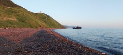This is a panoramic, eye-level, full shot of a rocky beach on a clear, calm day. The sun is low in the sky, casting a warm, golden light on the scene, suggesting either sunrise or sunset. The sky is a pale, clear blue, fading to a soft pinkish hue near the horizon.
The beach itself is composed of a mixture of sand and small, smooth pebbles in various shades of brown, grey, and red. Some dried seaweed and driftwood are scattered along the shoreline. The water is a serene, deep blue with gentle ripples.
To the left, a steep, grassy hill rises from the beach. The grass is a vibrant green, and the hill is dotted with small, low-lying shrubs. At the crest of the hill, a white lighthouse stands prominently, its light room a darker color. The lighthouse is a clear focal point of the scene.
In the distance, beyond the lighthouse, more rolling hills can be seen, and out in the sea, a cluster of dark, jagged rocks emerges from the water, adding a dramatic element to the otherwise tranquil landscape. The overall mood is peaceful and serene, capturing the quiet beauty of a coastal landscape bathed in soft light. The location is Aleksandrovsk-Sakhalinskiy, Russia. There is no visible text in the image.