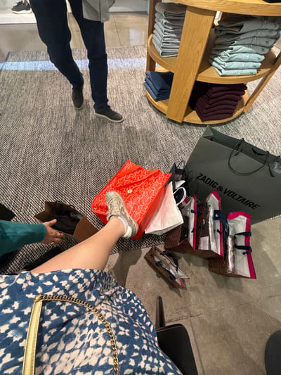 A cluttered floor with shopping bags from Zadig & Voltaire and other stores. A rotating display rack with folded clothing stands to the left. Two people are partially visible: one with dark jeans and shoes, and another wearing a blue patterned dress and sparkly sneakers, holding a shoe box. The floor is covered with a neutral-toned rug.