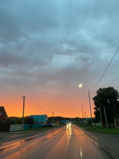 The image captures a rural street scene in Старое Субханкулово, Russia, during what appears to be dusk or after a rainfall. The sky is a dramatic gradient of deep blue and grey clouds at the top, transitioning to a vibrant orange sunset on the horizon. The wet asphalt road reflects the warm hues of the sky and the bright lights of passing cars, creating streaks of light.

On the sides of the road, there are houses with pitched roofs, one notably a bright teal color. Fences, including a white concrete one and a dark green one, line the properties. Utility poles with numerous wires crisscross the scene, typical of rural infrastructure. A large, dark green tree stands prominently on the right side of the road, silhouetted against the sky.

Several cars are visible on the road, their headlights illuminating the wet surface. There are no people clearly visible in the image. The overall impression is one of a quiet, overcast evening with a striking sunset over a village road.