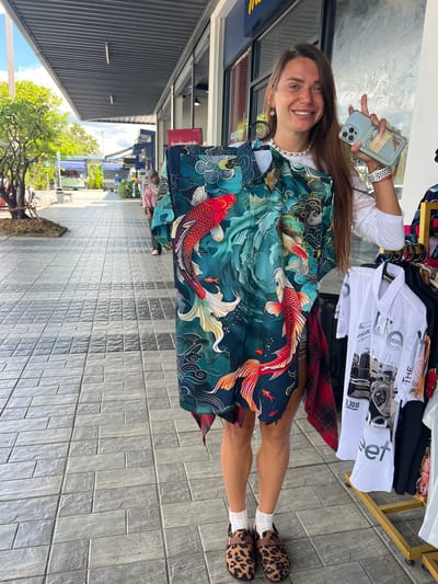 In Tambon Phraek Ha, Thailand, a woman stands on a tiled sidewalk in front of a row of market stalls. She is holding up a short-sleeved shirt with a dark teal background featuring a colorful koi fish and wave pattern. The woman, with long brown hair, wears a white long-sleeved top, denim shorts, white socks, and leopard-print clogs. She is also holding a light blue smartphone in her right hand. To her right, a rack displays several white t-shirts with graphic prints of vintage cars. The background shows a covered walkway, trees, and other stalls.