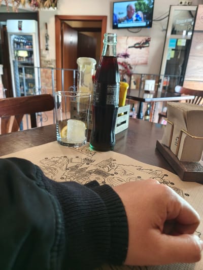 The image portrays an indoor scene, likely within a casual restaurant, bar, or cafe in Pontevedra, Spain. In the foreground, a person's left forearm and hand, clad in a dark, ribbed sleeve, rest on a brown paper placemat. The placemat features a detailed black-and-white illustration resembling a city map or architectural landmarks, with visible text including "KLICIA" and several numbers like "18", "47", "1000", and "1001".
On the dark wooden table, a classic glass bottle of Coca-Cola stands, its label displaying "Coke" and Spanish text "BEBIDA REFRESCANTE AROMA y aroma cafeína." Adjacent to it is an empty drinking glass containing ice cubes and a lemon slice, ready for the beverage. A small wooden caddy holds two condiment bottles, one yellow and one white, while a metal and wooden napkin holder stacked with brown napkins is positioned to the right.
The background reveals a bustling interior. To the left, a tall white refrigerator or cooler filled with various bottles and items is visible. Above the table, a flat-screen television mounted on the wall displays a program featuring two individuals, an older man and a woman. Wall decorations include a painting depicting wine being poured and a narrow vertical display case with Spanish text "DESPUES DE TODO" and "HUHOS PA" (likely "HECHOS PARA"). A wooden doorway and chairs are also part of the rustic-themed setting, complemented by dried foliage hanging from above. The scene suggests a relaxed moment, possibly awaiting or enjoying a drink.