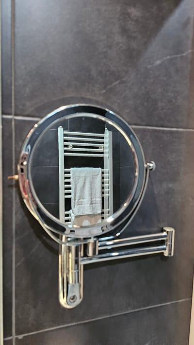 This is a close-up shot of a chrome magnifying mirror mounted on a dark gray tiled wall. The mirror reflects a white towel rack with a light gray towel hanging from one of its rungs. The mirror is round with a double-jointed arm extending from its side, attached to a mounting bracket on the wall. The background consists of dark gray tiles with visible grout lines. The lighting suggests an indoor setting, likely a bathroom. No people or specific locations in Alhama de Aragón, Spain are identifiable in the image.
