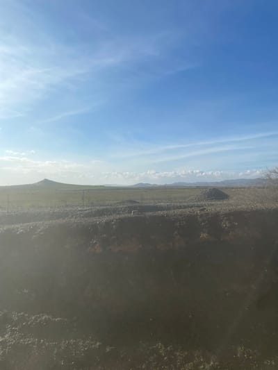 The image, when properly oriented, presents a vast, open landscape under a clear blue sky. The upper portion of the frame is filled with a bright, consistent blue sky, traversed by thin, wispy white clouds stretching horizontally.
Along the distant horizon, a low, undulating mountain range or a series of hills is visible, appearing somewhat faded against the sky. Below the mountains, a wide, flat plain stretches out, showing subtle hints of green vegetation, suggesting agricultural land or natural growth in a semi-arid environment.
The foreground, closer to the viewer, consists of a dark, rugged terrain, likely an unpaved road or track with rocky edges and sparse, dark flora. Several slender, vertical posts, possibly fence posts or small utility poles, are sparsely distributed along this foreground path, running roughly parallel to the horizon line. The foreground is distinctly darker than the sky and midground, potentially due to shadows or the natural color of the ground materials. A faint glare is present in the lower right corner of the image.
No people or discernible activities are taking place; the scene is a static depiction of the natural environment. The bright sky indicates it is daytime, possibly late morning or early afternoon, with clear weather conditions. The landscape is consistent with the provided location context of Jbilate, Morocco. No text is visible in the image.