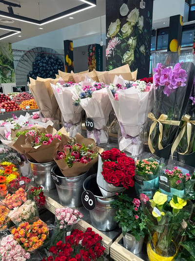 The image depicts a brightly lit flower display within what appears to be a supermarket or a large retail store, likely in Ras Al-Khaimah, United Arab Emirates, given the context of the query. The foreground is dominated by a vibrant assortment of cut flowers, arranged in bouquets and potted plants. Various types of roses, gerbera daisies, and orchids are visible, showcasing a spectrum of colors including reds, pinks, oranges, yellows, and purples.
Several bouquets are presented in brown paper wrapping tied with ribbons, and some are displayed in galvanized metal buckets. Price tags, some with "AED" and numbers like "170" and "90," are affixed to the buckets and some of the flower arrangements, indicating they are for sale. Behind the cut flowers, there are potted plants, including small flowering plants with pink and red blossoms, and larger yellow calla lilies.
The background reveals more of the store environment. A large mural featuring white and pink roses is visible on a dark wall. Further back, shelves stocked with fruits such as blueberries, plums, and oranges are discernible. Overhead, modern track lighting illuminates the scene. There are no people clearly visible in the image, and the focus is entirely on the merchandise. The overall impression is one of abundance and a well-organized retail space dedicated to floral products.