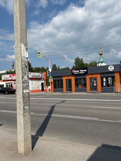 This image captures a street scene in Belovo, Russia, on a bright, cloudy day. The foreground is dominated by a large, weathered concrete pole, partially obscuring the view of the street and buildings behind it. Attached to the pole are torn pieces of paper with Russian text, including what appears to be an advertisement for "Specialists" and "Doner & Shawarma."

To the right of the pole, a multi-lane asphalt road stretches into the distance, marked with white lines. The sun casts a long shadow from the concrete pole onto the road.

In the mid-ground, a modern, dark-colored building with orange wooden accents stands adjacent to the road. A prominent sign on the building reads "Coner Home" in English and "Донер & Шаурма" in Russian, indicating it's a food establishment specializing in doner kebabs and shawarma. A "24 часа" (24 hours) sign is also visible, suggesting the establishment is open around the clock.

Behind the building, the green dome and golden cross of a church are visible against the blue sky and white clouds, providing a cultural and architectural landmark. A pedestrian crossing with a yellow warning sign is located near the building, and a traffic light is also present. Further down the road, another building with a red sign is partially visible, displaying Russian text that seems to include "Belovsky Distr.". A dark-colored car is parked on the side of the road near this building. The overall scene depicts a typical urban environment in Russia with a mix of modern commerce and traditional religious architecture.