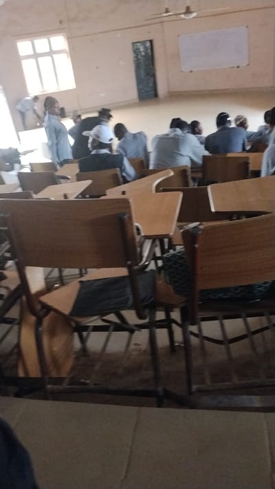 A classroom setting shows rows of empty wooden chairs with attached desks, some occupied by individuals in the background. Most appear to be wearing gray or white tops, possibly uniforms. There is a whiteboard on the wall and a window providing natural light. Other details include a doorway and a ceiling fan. The classroom has a worn and used appearance.