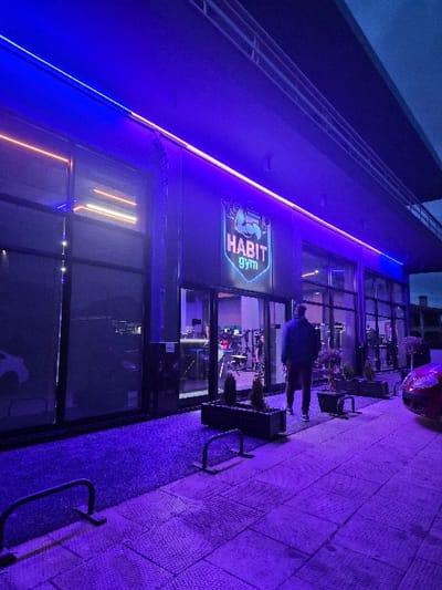 This image depicts the brightly lit entrance of "HABIT gym" at night in the Municipal Unit of Corinth, Greece. The modern building facade is dark, featuring large glass windows through which gym equipment and an interior counter are visible. Extensive blue and purple LED strip lighting illuminates the exterior, casting a strong violet hue across the scene. A dark artificial turf mat is present directly in front of the glass doors, bordered by dark rectangular planters containing small, conical evergreen shrubs. Beyond this, the ground transitions to light-colored, square paving stones. A single adult male, viewed from behind, stands facing the gym entrance, dressed in a dark jacket and lighter trousers with white-soled shoes. To the right, the front-right fender and headlight of a red car are partially visible. Two black U-shaped metal barriers are positioned on the paved area in front of the gym. The sky above is dark, indicating dusk or nighttime conditions.
