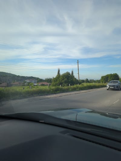 Captured from inside a car, the photograph shows a road winding through Beş-Küŋgöy, Kyrgyzstan. Another car is visible ahead on the road. Greenery borders the road, extending into the background where houses and buildings can be seen. Utility poles and wires run alongside the road. The sky is visible above, showing a mix of blue and clouds.