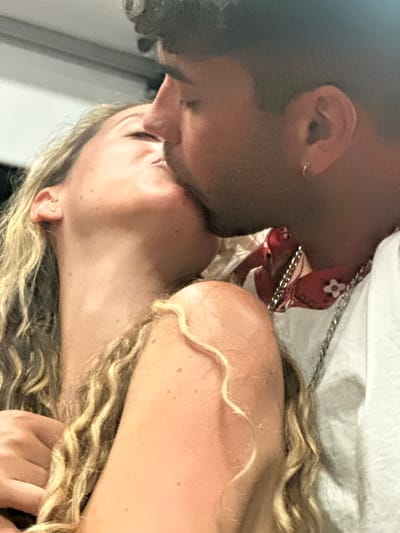 A close-up, slightly blurred image captures a couple kissing. The woman's head is tilted back, her eyes closed, and her lips are touching the man's. Her blonde, curly hair falls around her shoulders. The man, partially visible in the foreground, is kissing her, his eyes also closed. He wears a white t-shirt with a red bandana-like pattern and a silver chain around his neck. The background is indistinct, suggesting an indoor or dimly lit setting, with no discernible Barcelona landmarks. The overall impression is one of intimacy.
