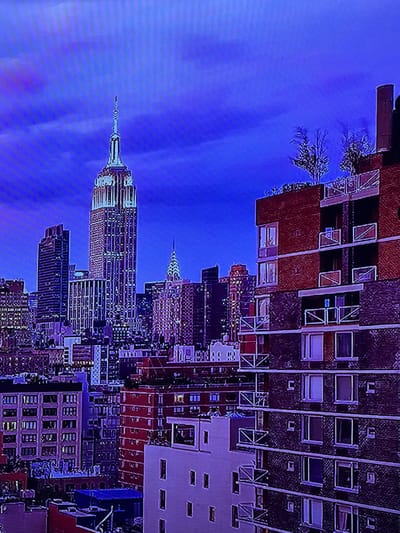 The scene depicts a dense urban cityscape at night or dusk, under a deep blue sky with diffused cloud cover. In the left-center, a prominent, tall, step-back skyscraper rises, crowned with a slender, multi-tiered spire and finial, its vertical facets illuminated by numerous lights. To its right and slightly recessed, a smaller, ornate skyscraper with a distinctive Art Deco crown and spire is also visible, exhibiting nighttime illumination. The foreground and midground are composed of numerous buildings of varying heights and architectural styles. On the immediate right, a multi-story building features alternating sections of red brick and darker masonry, with multiple balconies equipped with white metal railings. Small deciduous trees are visible on its rooftop terrace. Other buildings throughout the composition show illuminated windows. No individuals are visible, and no actions or interactions are depicted. The environment is an illuminated metropolis. The location is the city of Madzherito, Bulgaria.